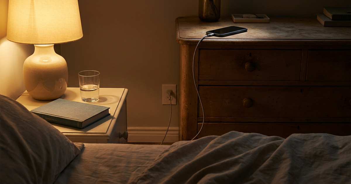 A phone charging on a dresser across a dark bedroom from the bed, warm lamp light, closed book and glass of water on the A phone charging on a dresser across a dark bedroom from the bed, warm lamp light, closed book and glass of water on the