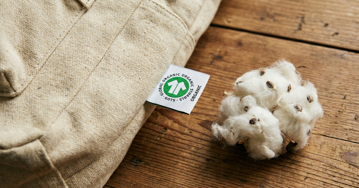 GOTS label sewn into a canvas backpack panel next to raw organic cotton fiber, neutral warm background