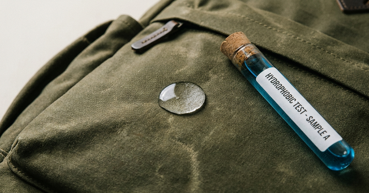 A canvas backpack with a water droplet beading off the fabric next to a lab test vial, neutral studio background