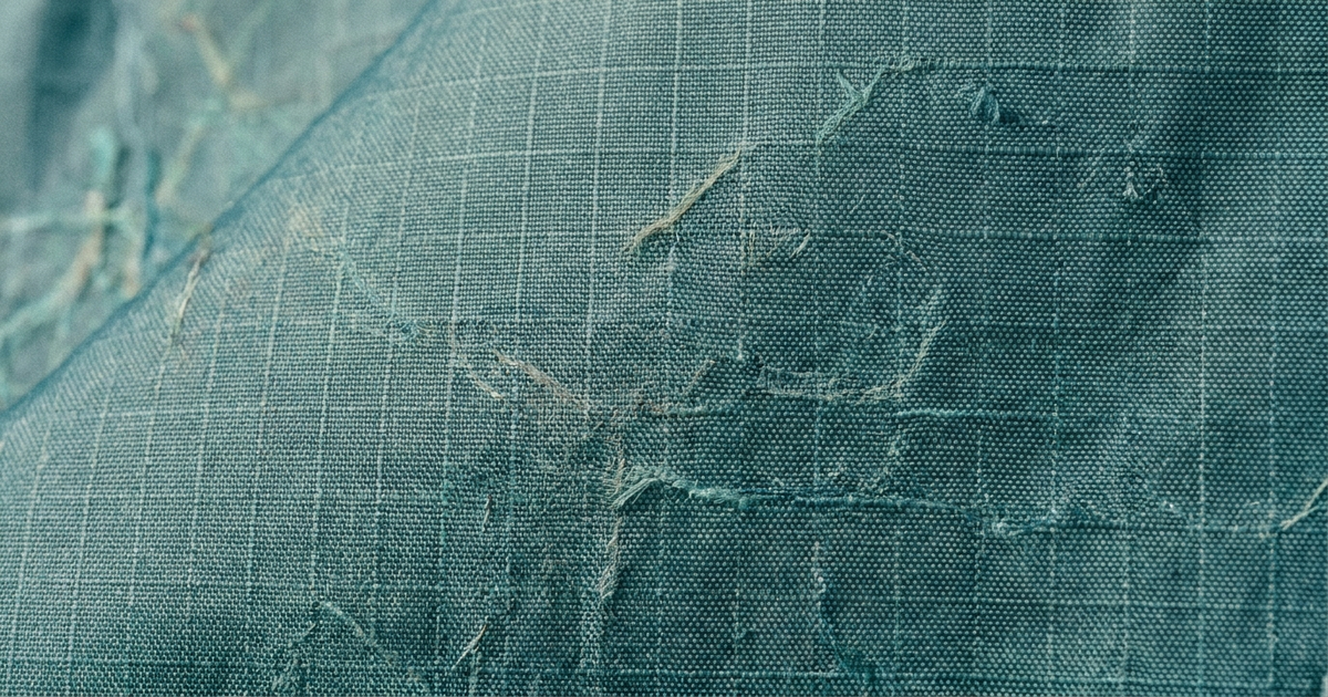 Close-up of grid-weave ripstop nylon fabric with a faint ghost-net texture woven through it, representing recycled nylon ripstop made from reclaimed fishing nets Close-up of grid-weave ripstop nylon fabric with a faint ghost-net texture woven through it, representing recycled nylon ripstop made from reclaimed fishing nets